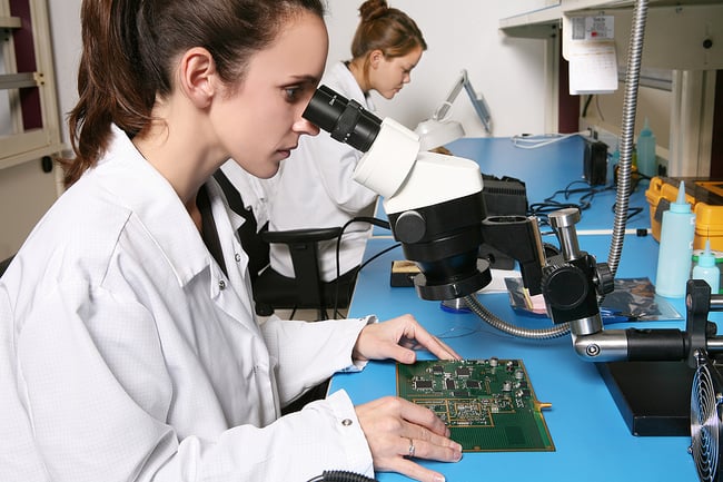 woman in an electronic manufacturing lab examining a printed circuit board with a microscope
